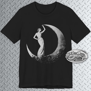 May include: Black t-shirt with a vintage-style graphic of a woman standing on a crescent moon. The woman is wearing a long dress and is reaching up to touch the moon. The design is in shades of white and grey.