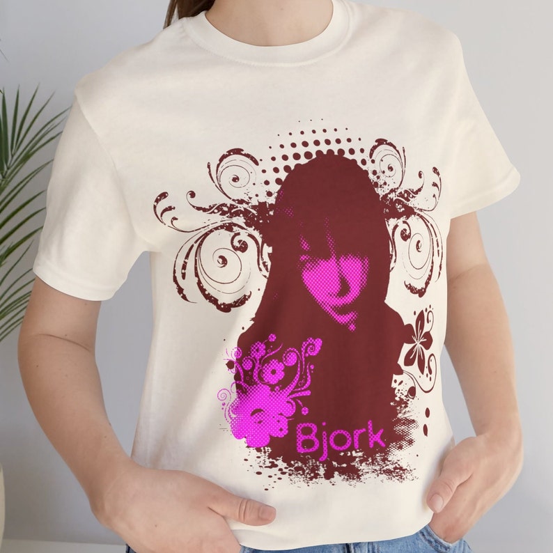 May include: White t-shirt with a maroon and pink graphic print of a woman's face with the text "Bjork" underneath.