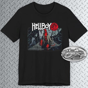 Op de afbeelding: Zwart T-shirt met een grafische print van Hellboy die op een begraafplaats staat. De grafiek is in rood, zwart en wit. De tekst "Hellboy" is in wit boven de grafiek gedrukt.