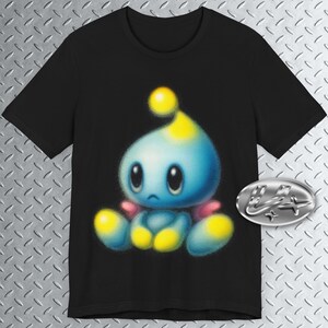 May include: Black t-shirt with a cartoon character design. The character has a blue body, yellow details, and a melancholic expression. A silver oval emblem is on the right. The design is centered on the shirt.