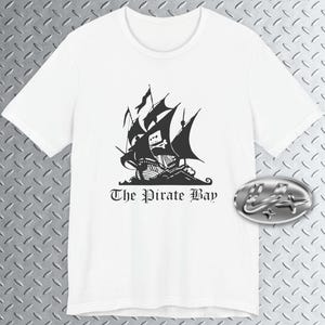 Può includere: Una t-shirt bianca a maniche corte con una grafica nera raffigurante un veliero con una bandiera con teschio e ossa incrociate. Sotto la nave, il testo The Pirate Bay è scritto in un carattere nero in stile gotico, centrato sul davanti.