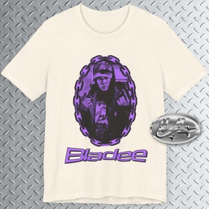 May include: A white t-shirt with a purple image of a person wearing a hat and a chain link oval around the image. The text "Bladee" is printed in purple below the image.