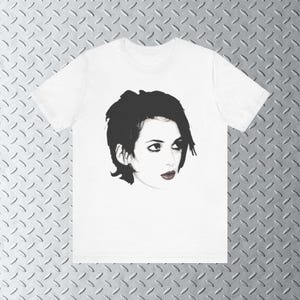 Pode incluir: Uma camiseta branca com uma imagem em preto e branco de uma mulher com cabelo curto e escuro. A mulher est&aacute; olhando para a direita da imagem.