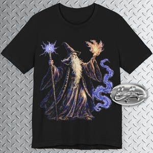 Może przedstawiać: Czarny T-shirt z grafiką przedstawiającą czarodzieja z długą białą brodą, trzymającego laskę i kulę ognia. Czarodziej jest ubrany w niebieską i fioletową szatę i otoczony fioletowym dymem.