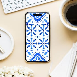 Op de afbeelding: Een smartphonehoesje met een doorzichtige rand en een blauw-wit bloemenpatroon. Het ontwerp toont gestileerde bloemen en geometrische vormen. De hoes ligt op een lichtbeige oppervlak, met een toetsenbord en een koffiekopje op de achtergrond.