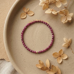 Puede incluir: Una delicada pulsera con pequeñas cuentas redondas de color rojo intenso. La pulsera tiene una pequeña cuenta dorada. Se exhibe en un plato de tono neutro, con flores secas esparcidas.