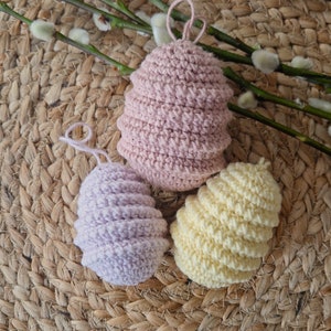 May include: Three crocheted Easter eggs in pink, purple, and yellow. The eggs are sitting on a woven straw mat.