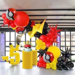 Peut inclure: Une arche de d&eacute;coration de f&ecirc;te d'anniversaire faite de ballons rouges, jaunes et noirs. L'arche est d&eacute;cor&eacute;e de ballons noirs en forme d'&eacute;toile et de ballons rouges et jaunes en forme d'&eacute;toile. Il y a aussi deux grands ballons rouges avec un motif de Pok&eacute; Ball. L'arche est install&eacute;e devant une fen&ecirc;tre avec vue sur une pelouse verte.