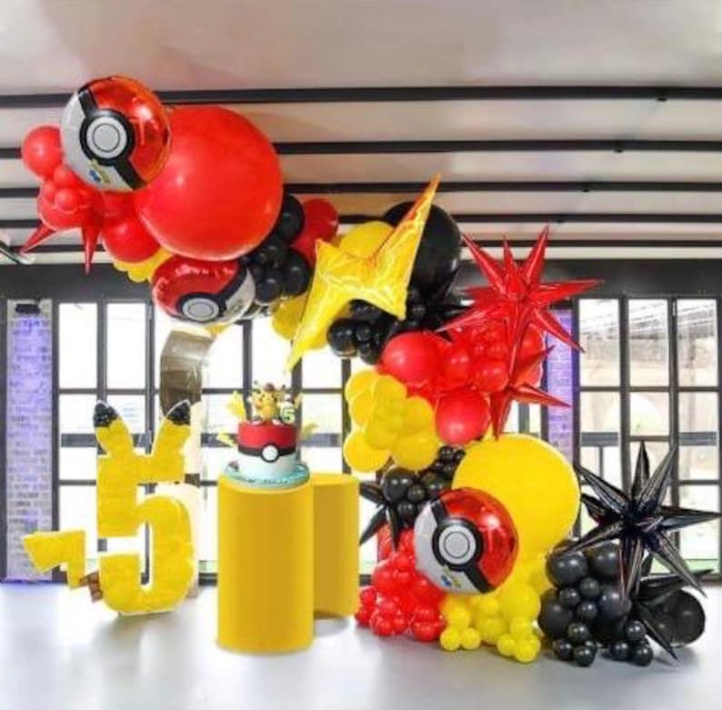 Peut inclure: Une arche de d&eacute;coration de f&ecirc;te d'anniversaire faite de ballons rouges, jaunes et noirs. L'arche est d&eacute;cor&eacute;e de ballons noirs en forme d'&eacute;toile et de ballons rouges et jaunes en forme d'&eacute;toile. Il y a aussi deux grands ballons rouges avec un motif de Pok&eacute; Ball. L'arche est install&eacute;e devant une fen&ecirc;tre avec vue sur une pelouse verte.