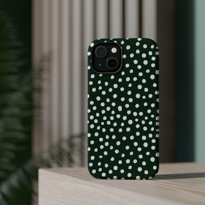 K&ouml;nnte beinhalten: Dunkelgr&uuml;ne Handyh&uuml;lle mit wei&szlig;em Polka-Dot-Muster. Die H&uuml;lle hat einen schwarzen Kameraobjektiv-Ausschnitt und eine glatte, gl&auml;nzende Oberfl&auml;che. Die Handyh&uuml;lle liegt auf einer hellen Holzoberfl&auml;che.