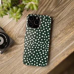 K&ouml;nnte beinhalten: Dunkelgr&uuml;ne Handyh&uuml;lle mit wei&szlig;em Polka-Dot-Muster. Die H&uuml;lle liegt auf einer Holzoberfl&auml;che, mit einer Kamera und einer Pflanze im Hintergrund. Die Handyh&uuml;lle hat eine gl&auml;nzende Oberfl&auml;che und ein sch&uuml;tzendes Design.