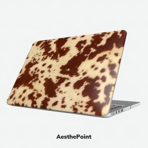Può includere: Computer portatile con un motivo a pelle di mucca sul coperchio. Il design presenta uno sfondo beige con macchie marroni irregolari, che imitano l'aspetto della pelle animale. Il computer portatile è aperto, rivelando la tastiera e lo schermo.