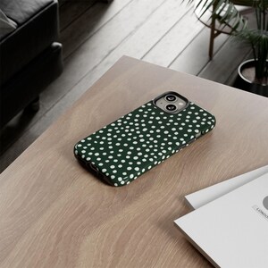 K&ouml;nnte beinhalten: Dunkelgr&uuml;ne Handyh&uuml;lle mit wei&szlig;em Polka-Dot-Muster. Die H&uuml;lle ist rechteckig und hat einen Kameraausschnitt. Sie liegt auf einer hellbraunen Holzoberfl&auml;che.