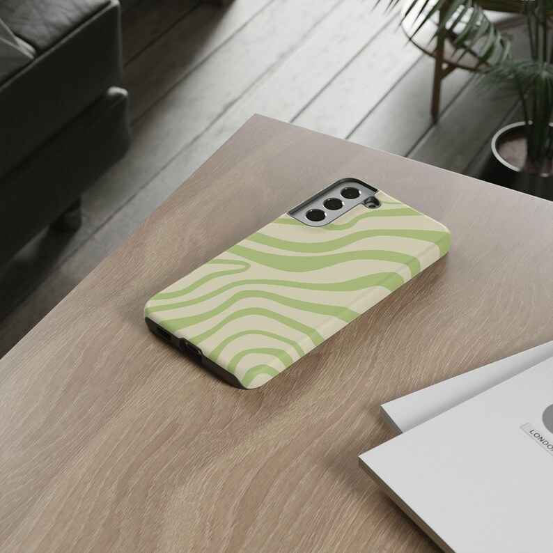K&ouml;nnte beinhalten: Eine Smartphone-H&uuml;lle mit einem wellenf&ouml;rmigen Muster in Hellgr&uuml;n und Creme. Die H&uuml;lle liegt auf einer Holzoberfl&auml;che und zeigt ihr Design und ihre Textur. Die H&uuml;lle ist f&uuml;r ein Smartphone konzipiert.