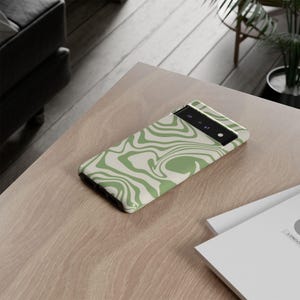 Peut inclure: Une coque de t&eacute;l&eacute;phone avec un motif tourbillonnant vert et cr&egrave;me. La coque rectangulaire pr&eacute;sente une d&eacute;coupe pour l'appareil photo en haut. Elle est pos&eacute;e sur une table en bois brun clair. Un accessoire moderne.