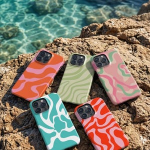 Puede incluir: Cinco fundas de teléfono coloridas con diseños ondulados abstractos. Las fundas presentan colores vibrantes como naranja, rosa, verde y turquesa. Las fundas se muestran sobre una superficie rocosa cerca del mar.