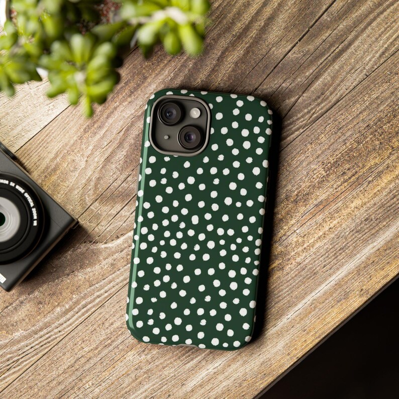 K&ouml;nnte beinhalten: Dunkelgr&uuml;ne Handyh&uuml;lle mit wei&szlig;em Polka-Dot-Muster. Die H&uuml;lle liegt auf einer Holzoberfl&auml;che, neben einer schwarzen Kamera und einer gr&uuml;nen Pflanze. Die Handyh&uuml;lle hat ein gl&auml;nzendes Finish.