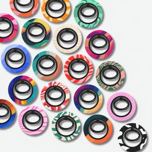 Può includere: Una collezione di anelli per telefono colorati con vari motivi. Ogni anello ha un anello di metallo nero attaccato a una base circolare. Le basi presentano disegni in tonalità di arancione, rosa, verde, blu e bianco, con motivi astratti e ondulati.