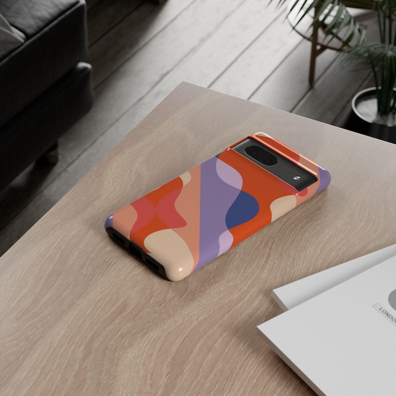 Peut inclure: Coque de t&eacute;l&eacute;phone avec un motif abstrait color&eacute;. Le design comprend des formes orange, rose, violet et bleu. La coque est pos&eacute;e sur une surface en bois clair.