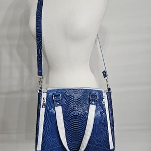 May include: A blue faux snakeskin tote bag with white accents. The bag has a rectangular shape with a zipper closure and a detachable shoulder strap. The bag is displayed on a mannequin.