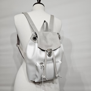 May include: A white and gray backpack with a flap closure and drawstring. The backpack has a front pocket with a zipper and adjustable straps. A blank tag hangs from the bag.
