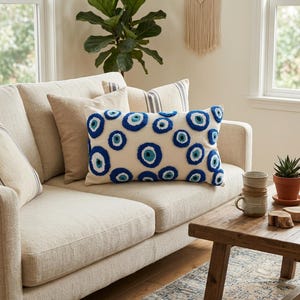 May include: A rectangular throw pillow with a cream-colored fabric base featuring a repeating pattern of tufted, circular evil eye motifs in shades of dark blue, light blue, and white, creating a textured, raised surface across the front.