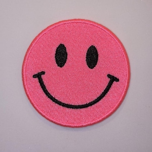May include: A circular embroidered patch featuring a bright pink background with a black stitched smiley face design. The patch has two black oval eyes and a wide black curved mouth, all rendered in a dense thread texture with a finished border.