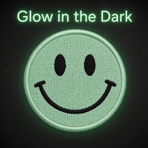 May include: A circular embroidered patch featuring a light green smiley face with black eyes and a black curved mouth. Above the patch, the text Glow in the Dark is written in a matching light green font against a dark background.