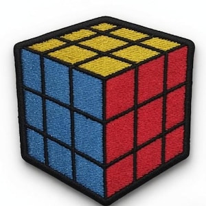 Op de afbeelding: Een geborduurde patch in de vorm van een driedimensionale kubuspuzzel. De bovenkant is geel, de linkerkant is blauw en de rechterkant is rood. Elk vlak is verdeeld in een drie-bij-drie raster door dikke zwarte geborduurde lijnen.