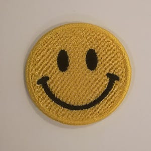 May include: A yellow embroidered patch featuring a classic smiley face design. The patch has a circular shape with a yellow background and black eyes and a curved mouth. The patch is likely for clothing or accessories.
