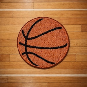 May include: Round, orange basketball rug with black lines, resembling a basketball. The rug is placed on a wooden floor with a white line, creating a sports-themed aesthetic. The rug's texture is visible, adding depth to the design.