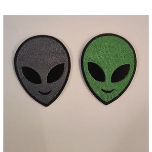 Op de afbeelding: Twee geborduurde alien patches. Eén is grijs met zwarte ogen en een glimlach, de andere is groen met zwarte ogen en een glimlach. Beide hebben een zwarte rand en staan tegen een witte achtergrond.