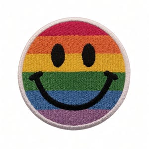 May include: A circular embroidered patch featuring a horizontal rainbow stripe pattern in red, orange, yellow, green, blue, and purple. The patch displays a black stitched smiley face with two oval eyes and a wide, curved mouth centered on the stripes.