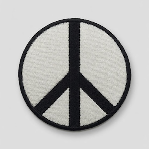 Puede incluir: Un parche bordado circular que presenta un símbolo de la paz negro centrado sobre un fondo blanco. El diseño está encerrado por un grueso borde cosido en negro, creando un elemento gráfico de alto contraste adecuado para aplicar sobre diversas telas.