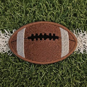 May include: A brown football patch with white stripes and black stitching, set against a backdrop of green turf and a white yard line. The patch is oval-shaped and detailed.