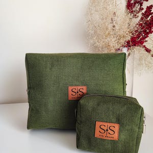 Könnte beinhalten: Zwei grüne Reißverschlusstaschen mit braunen Lederpatches, auf denen "SS SISI BRAND" steht. Die größere Tasche befindet sich hinter der kleineren.