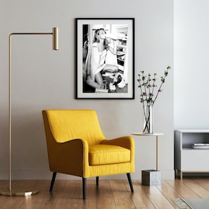 May include: A black and white photograph of a woman in a room, framed in a black frame. The woman is wearing a white shirt and is holding a glass of water. The photograph is hanging on a white wall above a yellow armchair.