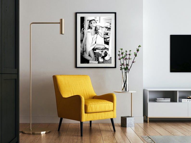 May include: A black and white photograph of a woman in a room, framed in a black frame. The woman is wearing a white shirt and is holding a glass of water. The photograph is hanging on a white wall above a yellow armchair.