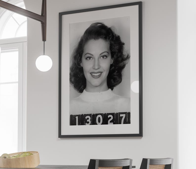 May include: Black and white photograph of a woman with dark hair, wearing a white shirt. The photo is framed in black and has the number "13027" printed at the bottom.