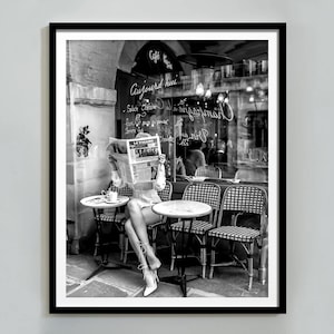 Puede incluir: Una fotografía en blanco y negro de una mujer sentada en una mesa de café en París, Francia. Está leyendo un periódico y tiene una taza de café delante de ella. El café se llama "Cafe des Arts" y el letrero dice "Aujourd'hui huitre et champagne".