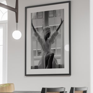 May include: Black and white photograph of a person wearing a white robe and a towel on their head, standing in front of a window with their arms raised.