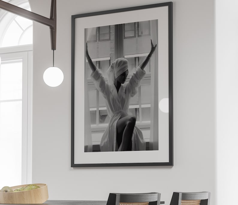 May include: Black and white photograph of a person wearing a white robe and a towel on their head, standing in front of a window with their arms raised.