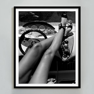 May include: Black and white photograph of a person's legs in a boat. The person is wearing high heels and is sitting in the driver's seat. The dashboard of the boat is visible, with gauges and a steering wheel.