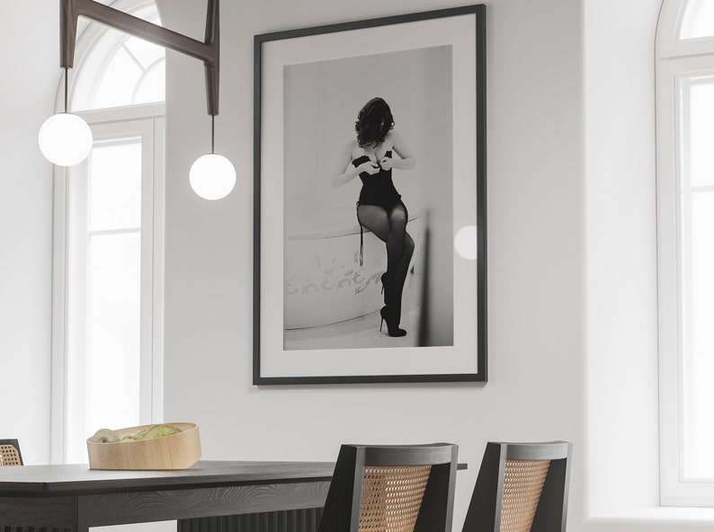 K&ouml;nnte beinhalten: Gerahmte Schwarz-Wei&szlig;-Fotografie einer Person in Dessous, die an einer wei&szlig;en Wand h&auml;ngt. Das Foto ist der Mittelpunkt eines Esszimmers, mit einem dunklen Holztisch und St&uuml;hlen im Vordergrund.