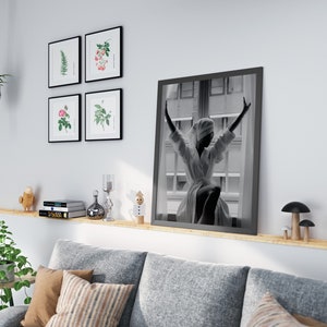 May include: A living room interior with a gray sofa, a wooden shelf with books, a small potted plant, and a black framed photo of a woman in a white robe with her arms raised.