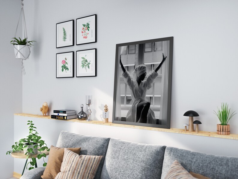May include: A living room interior with a gray sofa, a wooden shelf with books, a small potted plant, and a black framed photo of a woman in a white robe with her arms raised.
