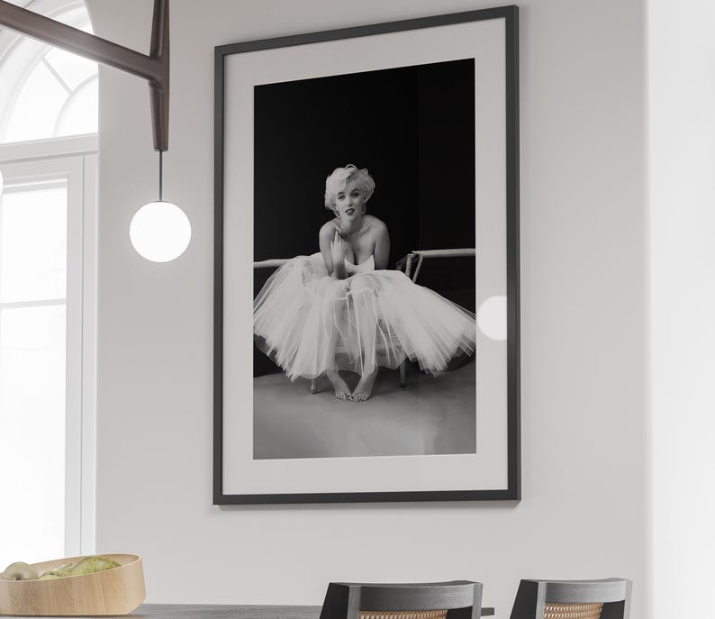 May include: Black and white photograph of a woman wearing a white tulle skirt and sitting on a chair. The woman is looking at the camera and smiling.