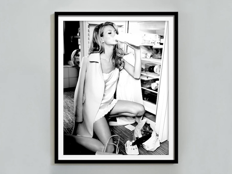 May include: A black and white photograph of a woman in a white coat and dress, standing in front of a refrigerator, drinking from a bottle.