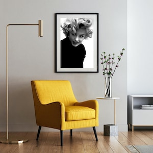 Peut inclure: Une photographie en noir et blanc d'une femme aux cheveux blonds, portant un haut noir, encadr&eacute;e dans un cadre noir. La photo est accroch&eacute;e &agrave; un mur blanc au-dessus d'un fauteuil jaune.