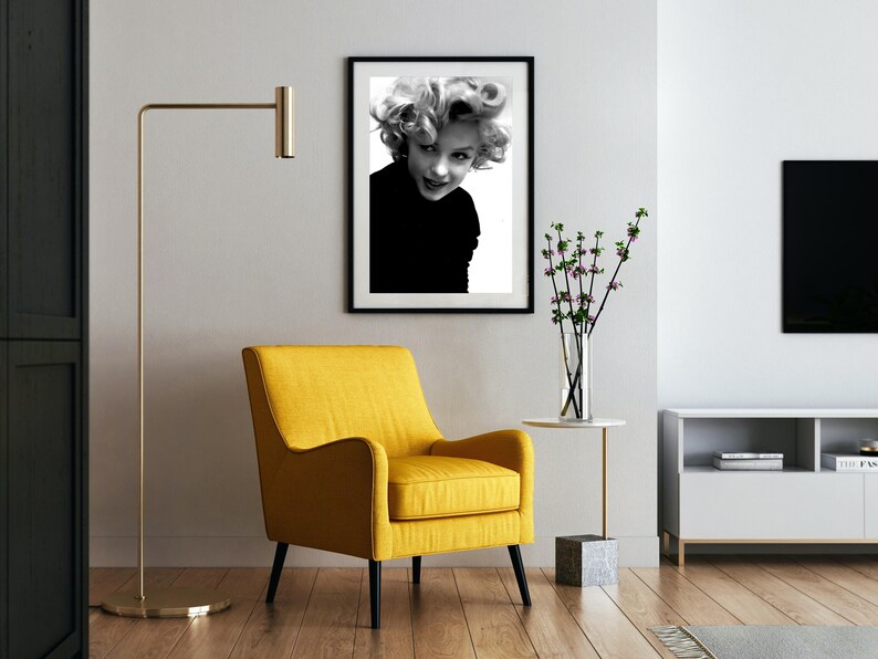 Peut inclure: Une photographie en noir et blanc d'une femme aux cheveux blonds, portant un haut noir, encadr&eacute;e dans un cadre noir. La photo est accroch&eacute;e &agrave; un mur blanc au-dessus d'un fauteuil jaune.